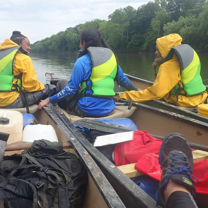 The image shows three people in a canoe on a river, likely engaged in a recreational activity. They are wearing life jackets, suggesting a focus on safety. The canoe is filled with gear, including containers and bags, indicating a possible multi-day trip. The surrounding environment features lush green trees lining the riverbanks, creating a natural and serene backdrop for their adventure.